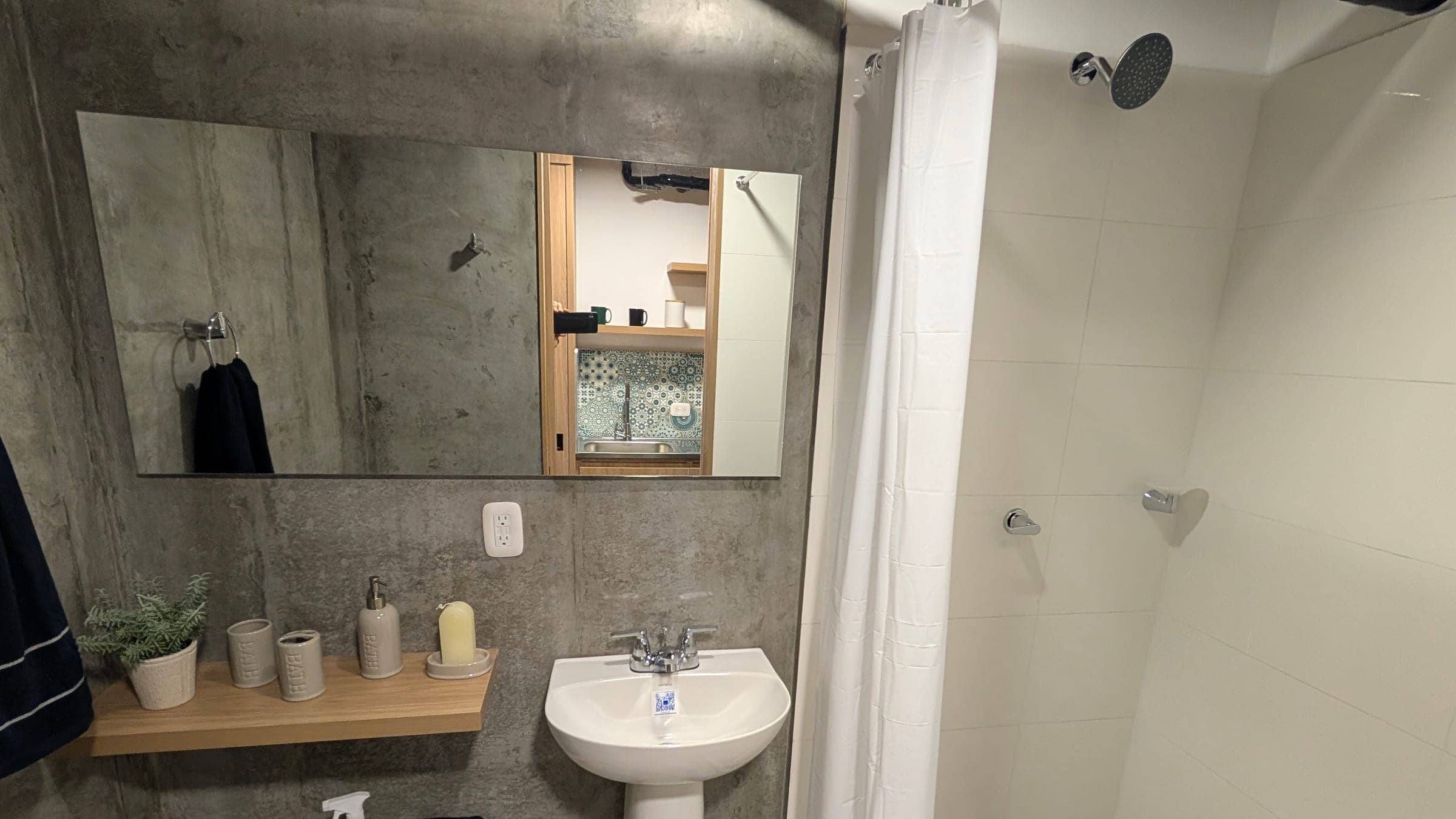 Baño con lavamanos blanco de pedestal, espejo horizontal grande sobre muro de concreto, dos repisas de madera flotantes, ducha tipo lluvia con cortina blanca y piso de baldosa hidráulica de colores.
