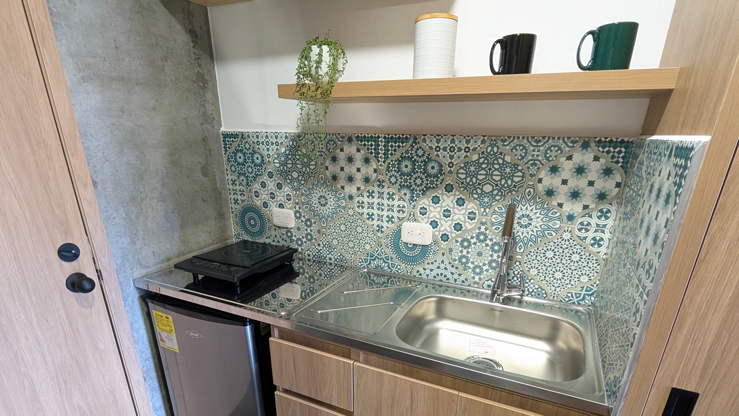 Cocineta con mueble bajo de madera clara, nevera, estufa de inducción de un puesto sobre mesón de acero y poceta de acero inoxidable con grifería alta. Pared de baldosa turquesa estampada y una repisa de madera con tazas.