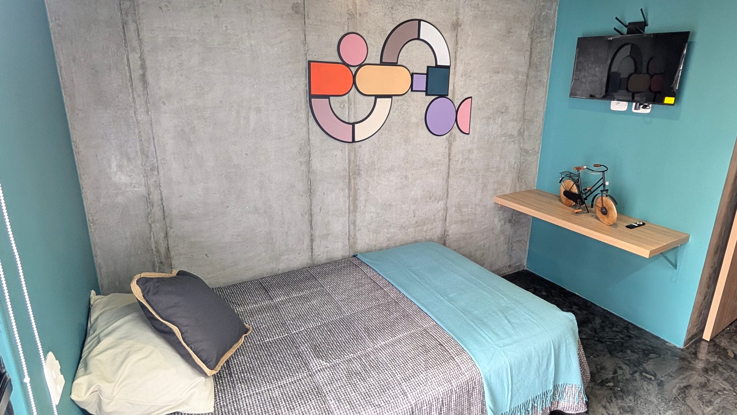 Cama semidoble tendida contra un muro de concreto a la vista con un mural de círculos y rectángulos naranjas, rosados y lilas. Paredes turquesa, televisor plano en la pared con el router de internet encima y una repisa de madera al lado.