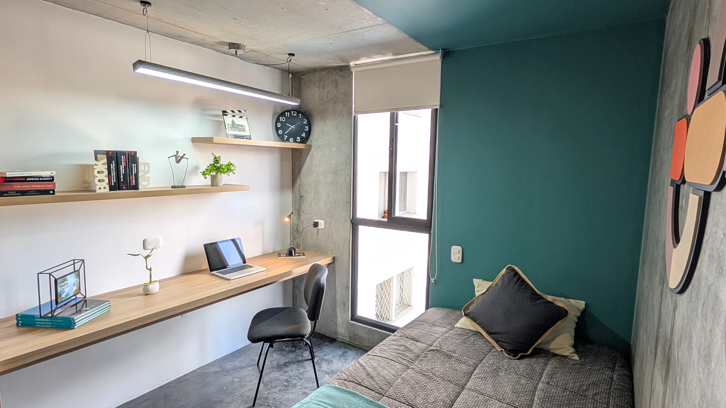 Escritorio de madera corrido a lo largo de la pared, bajo una repisa larga con libros, marcos y plantas. Silla de espaldar negro, lámpara lineal encendida sobre la mesa, ventana alta con persiana y, al fondo, la cama del apartaestudio.